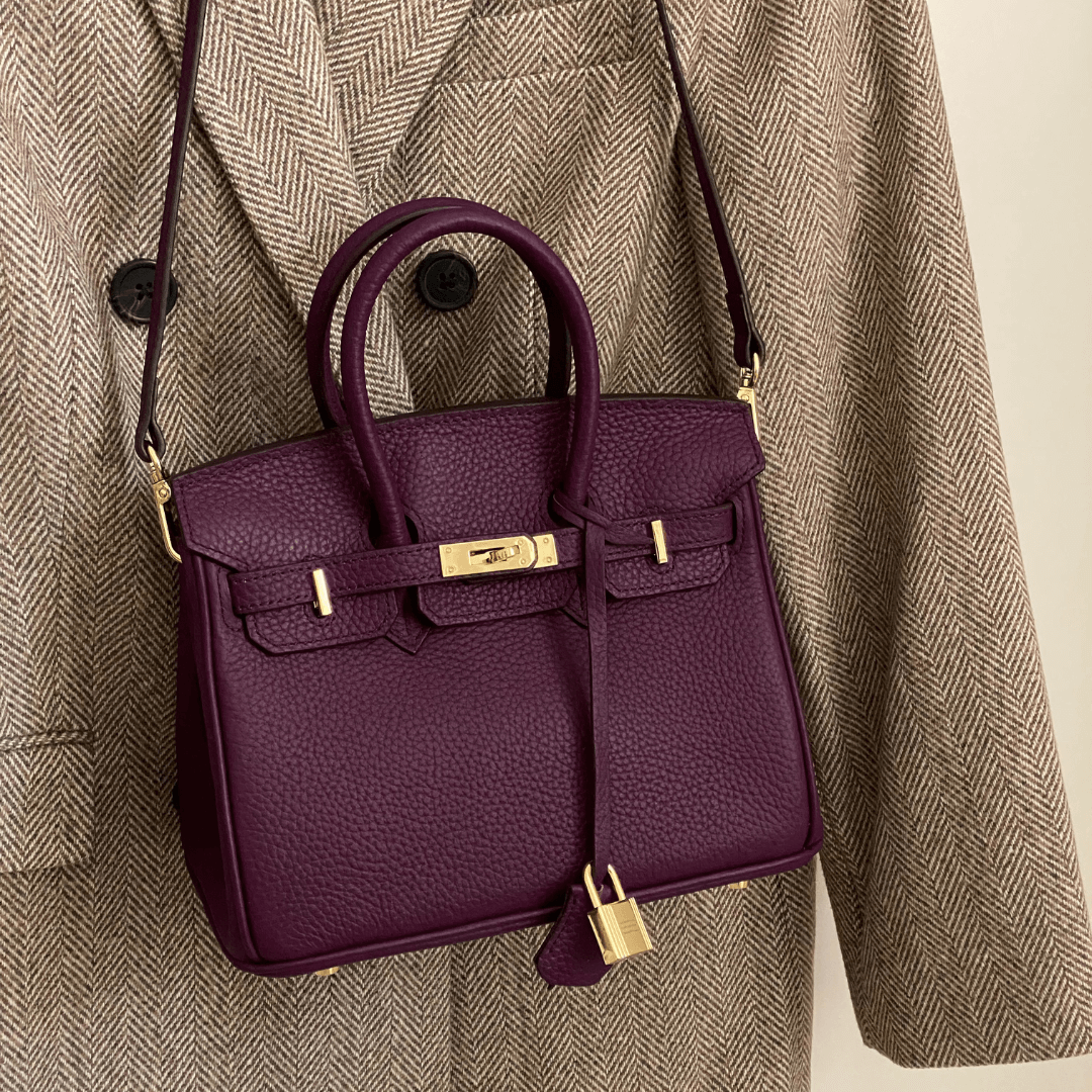 Lila Designer-Handtasche aus Leder mit goldenen Details, hängend an einem beigen Mantel. Luxuriöse Modeaccessoires, stilvoll und elegant.