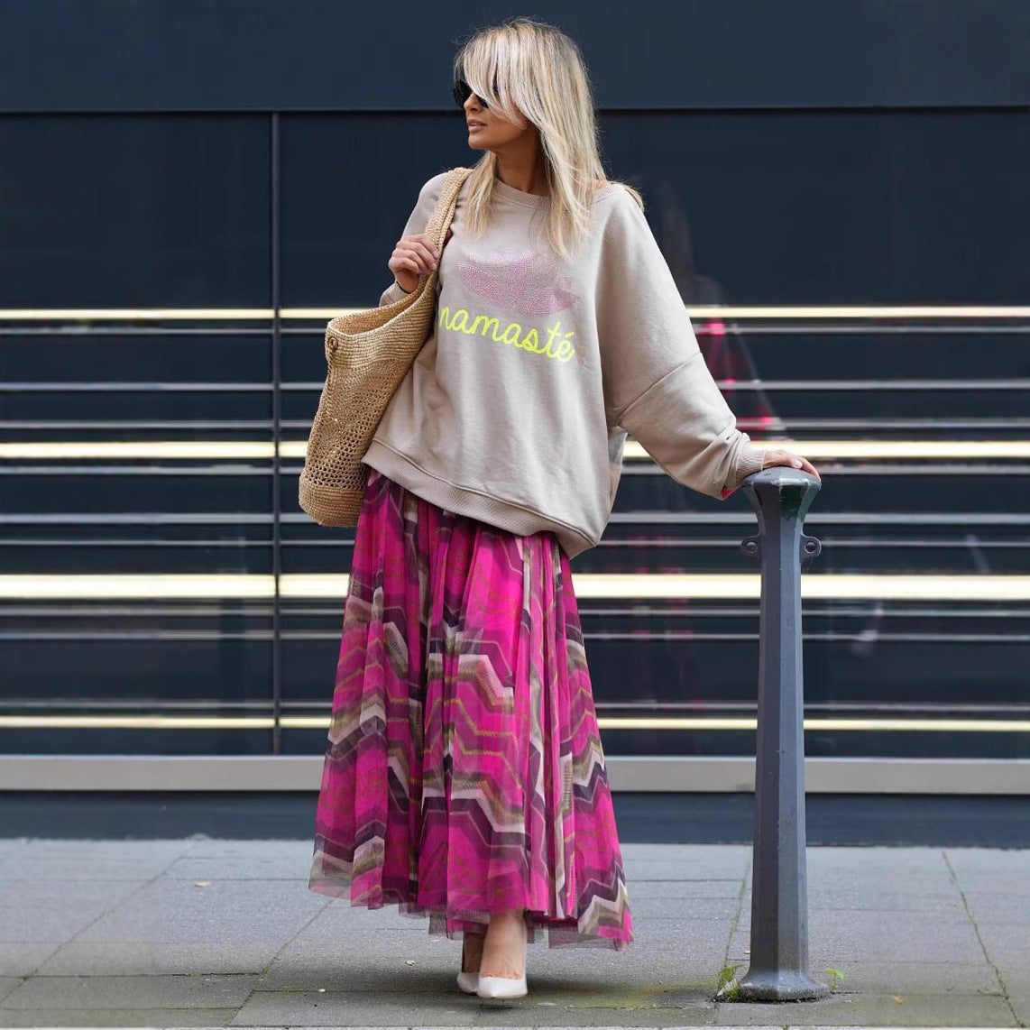 Frau in beiger "Namaste"-Sweatshirt und pinkem Maxirock, trägt Strohtasche, steht vor modernem Gebäude. Streetstyle, Mode, urbaner Look.