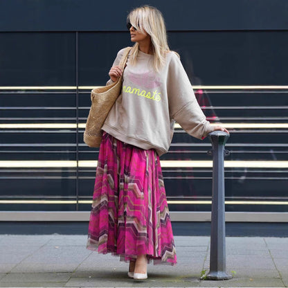 Frau in beiger "Namaste"-Sweatshirt und pinkem Maxirock, trägt Strohtasche, steht vor modernem Gebäude. Streetstyle, Mode, urbaner Look.