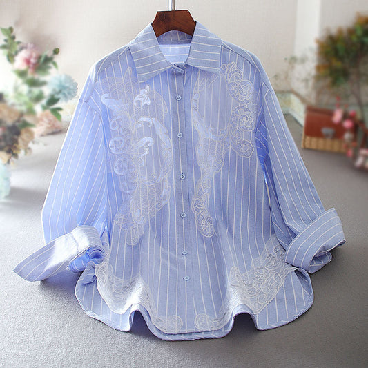 Hellblaues gestreiftes Hemd mit floralen Stickereien, langärmlig, auf einem Kleiderbügel. Elegante Herrenmode, ideal für formelle Anlässe.