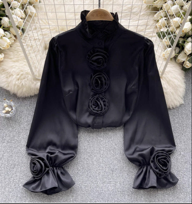 Schwarze Satinbluse mit Rosenverzierungen, langen Ärmeln und Stehkragen, elegant drapiert auf grauem Hintergrund, umgeben von weißen Blumen.