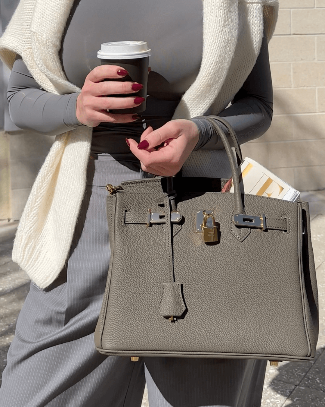 Frau mit grauer Designer-Handtasche und Kaffeebecher, trägt grauen Pullover und Schal. Mode, Accessoires, Streetstyle, Herbstmode.