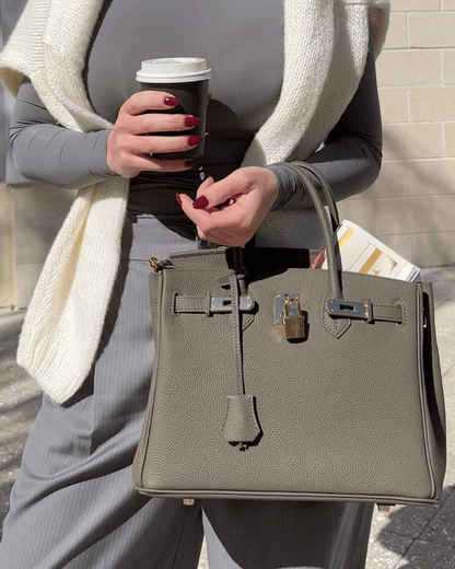 Frau mit grauer Designer-Handtasche und Kaffeebecher, trägt grauen Pullover und Schal. Mode, Accessoires, Streetstyle, Herbstmode.