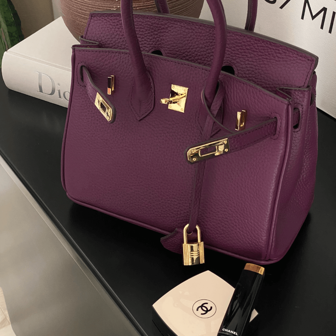 Luxuriöse lila Designer-Handtasche aus Leder mit goldenen Beschlägen, platziert neben Chanel-Make-up auf einem schwarzen Tisch.