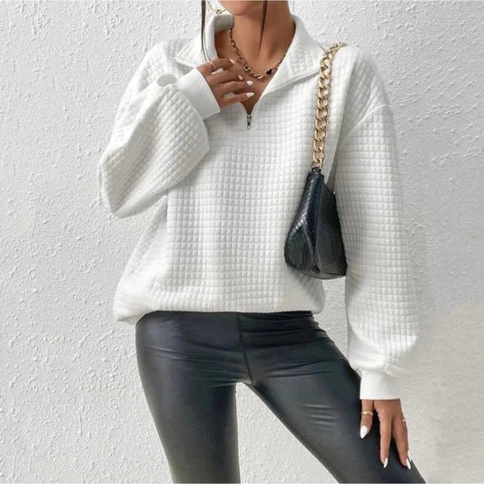 Weiße gesteppte Pullover mit Reißverschluss, kombiniert mit schwarzer Lederhose und schwarzer Umhängetasche mit goldener Kette, vor weißer Wand.