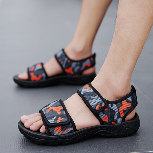 Person trägt Camouflage-Sandalen mit Klettverschluss auf grauem Boden. Bequeme, modische Sommerschuhe für Herren. Outdoor-Sandalen in Schwarz und Rot.