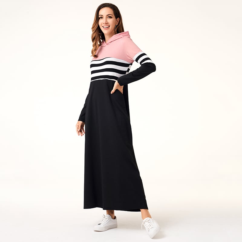 Frau in langem, gestreiftem Kapuzenkleid in Schwarz, Weiß und Rosa, lächelt und trägt weiße Sneaker. Modetrend, bequemes Damenoutfit.