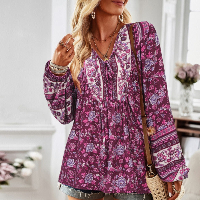 Frau in lila geblümter Bluse mit langen Ärmeln, Jeansshorts und Schmuck, steht in elegantem Raum mit Blumen im Hintergrund. Boho-Chic Mode.