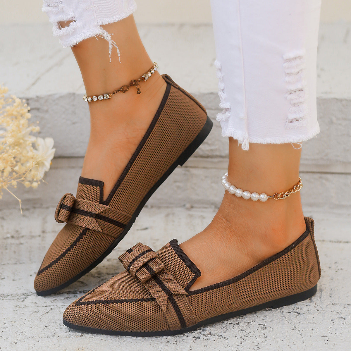 Braune Damen-Loafer mit Schleife, getragen mit weißen, zerrissenen Jeans und Perlenknöchelketten. Elegante, modische Schuhe für den Alltag.