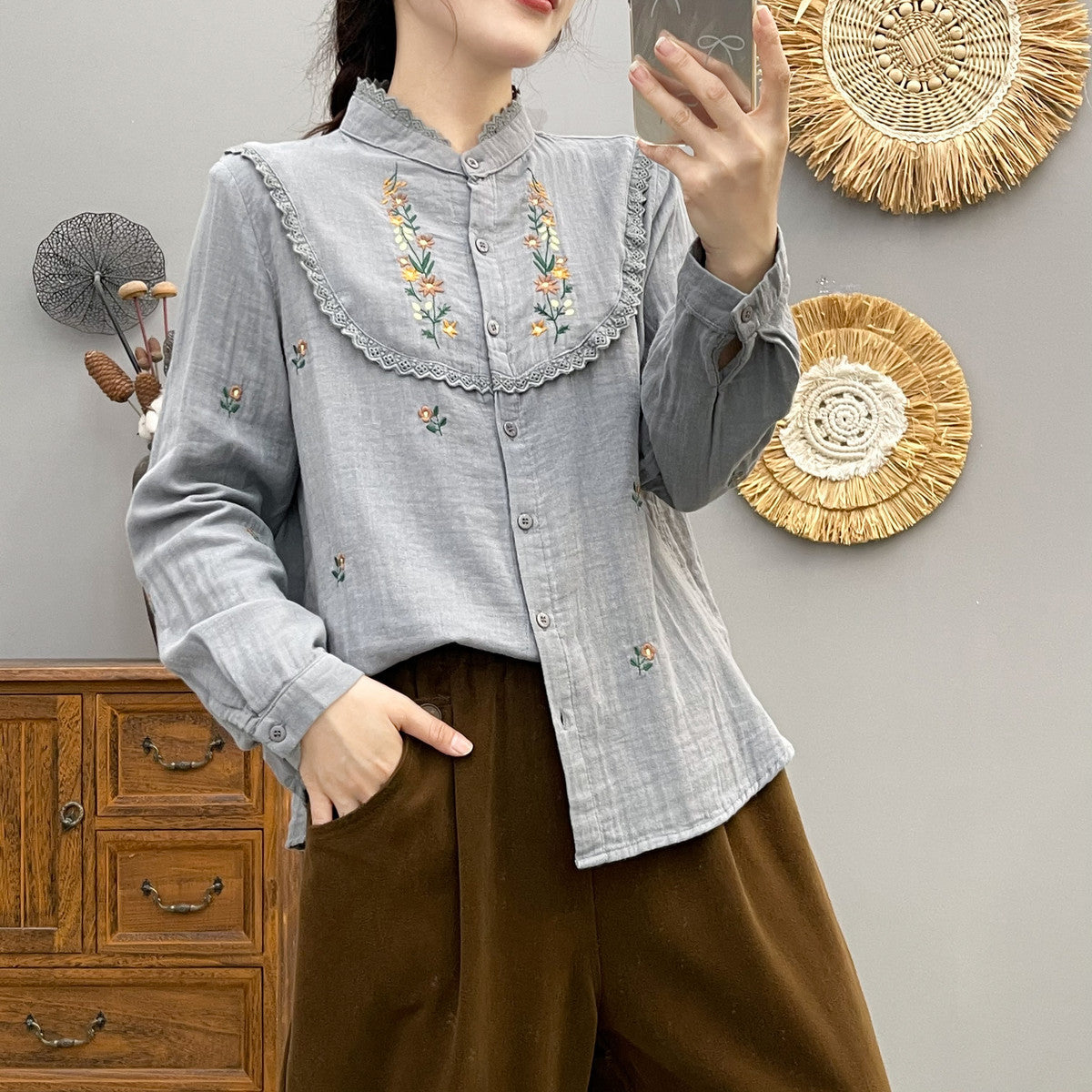 Frau in grauer Bluse mit Blumenstickerei, posiert vor Holzmöbeln und Wanddekorationen. Modetrend, Damenmode, Stickerei-Design.