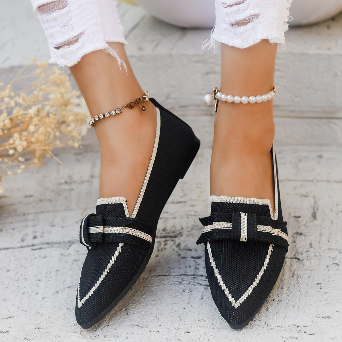 Damenfüße in schwarzen Loafers mit Schleifen, weiße Jeans, Perlen- und Gliederfußkettchen, stilvoller Look, modische Schuhe, Sommermode.
