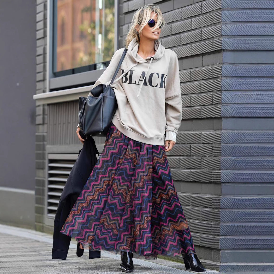 Frau in grauem Hoodie mit "Black"-Aufdruck, bunter Maxirock, schwarze Stiefel, Sonnenbrille, schwarze Tasche, steht vor grauer Backsteinwand.