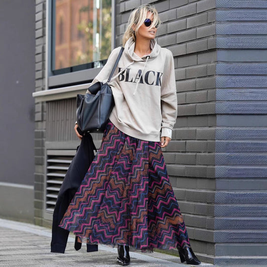 Frau in grauem Hoodie mit "Black"-Aufdruck, bunter Maxirock, schwarze Stiefel, Sonnenbrille, schwarze Tasche, steht vor grauer Backsteinwand.