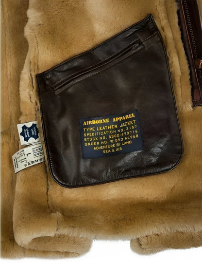 Braune Lederjacke mit Etikett "Airborne Apparel", auf beigem Fellhintergrund. Detailansicht der Tasche mit blauer und gelber Aufschrift.
