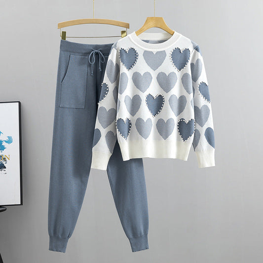 Grauer Jogginganzug mit Herzmuster-Pullover, hängend an Holzbügeln vor grauem Hintergrund. Bequeme Freizeitmode, stilvoll und modern.