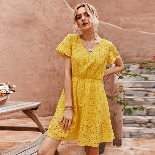 Frau in gelbem Sommerkleid mit Spitzenmuster, steht vor einer Terrakotta-Wand. Perfektes Outfit für warme Tage, modisch und elegant.