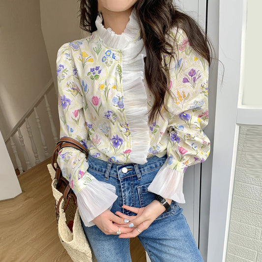 Frau in geblümter Bluse mit Rüschen und blauen Jeans, hält eine Strohtasche. Modetrend, Frühlingsoutfit, feminine Kleidung, Blumenmuster.