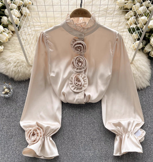 Seidenbluse mit langen Ärmeln und Rosendetails, elegant drapiert auf grauem Teppich, umgeben von weißen Rosen. Perfekt für stilvolle Anlässe.