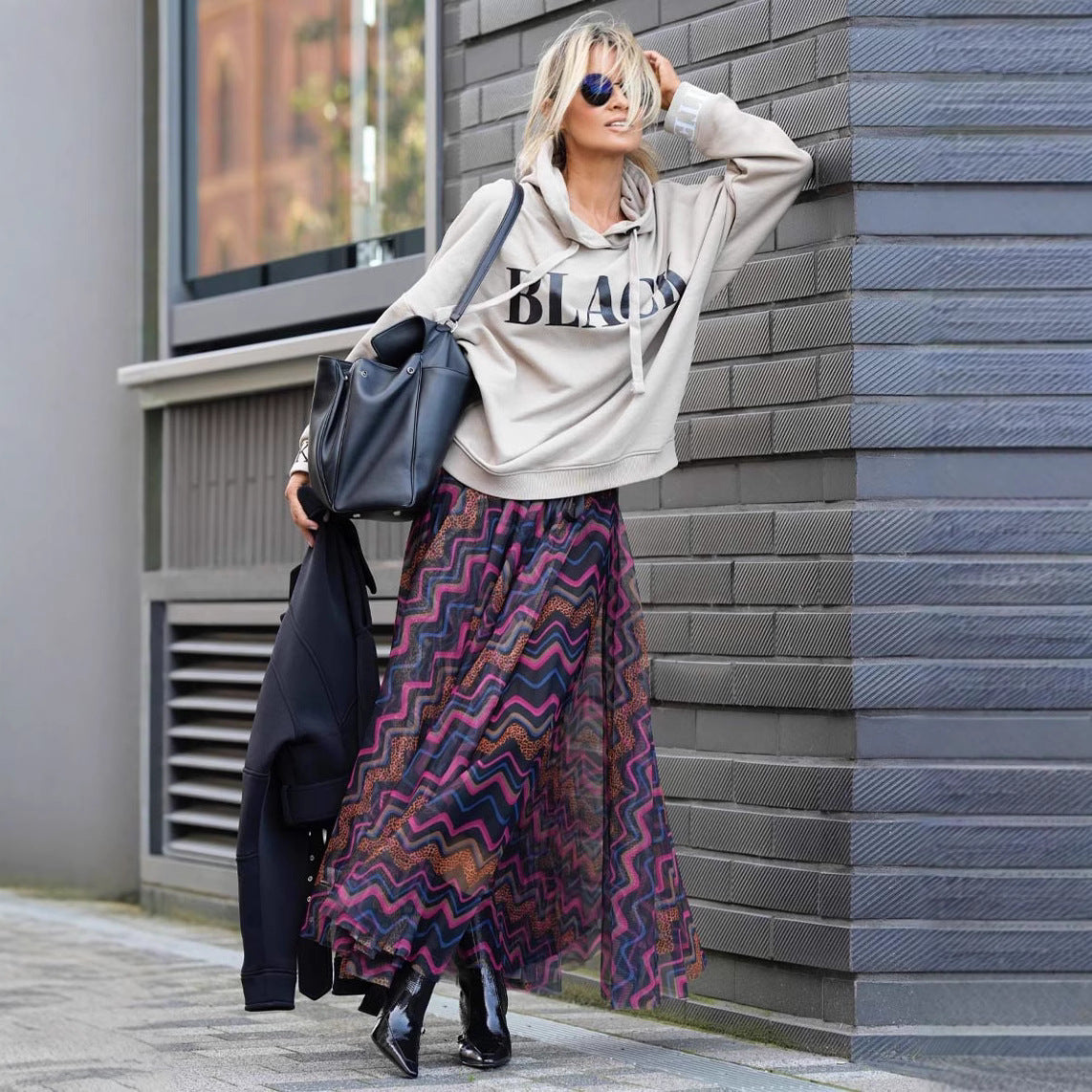 Blonde Frau in grauem Hoodie mit "Black"-Aufdruck, bunter Maxirock, schwarze Stiefel, Sonnenbrille, schwarze Tasche, lehnt an grauer Wand.