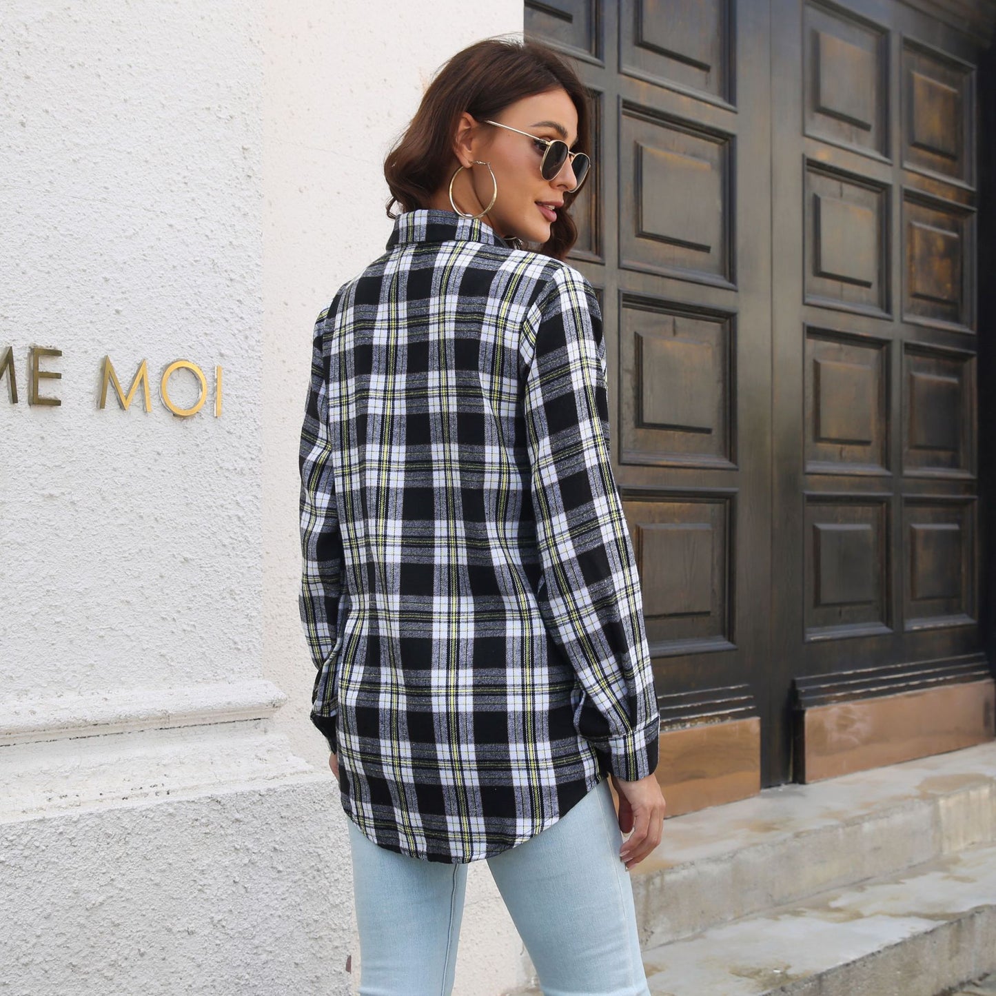 Frau in kariertem Hemd und Jeans vor Holztür, seitlich stehend. Modetrend, lässiger Stil, Herbstmode, urbane Kleidung, Streetwear.