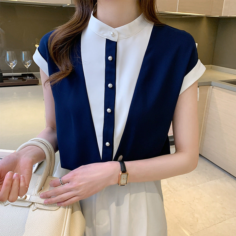 Frau in eleganter, zweifarbiger Bluse mit Perlenknöpfen, hält weiße Handtasche. Moderne Kücheneinrichtung im Hintergrund.