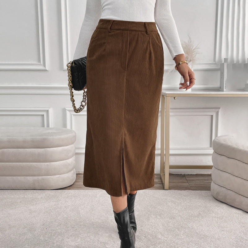 Frau in weißem Pullover und braunem Cordrock, kombiniert mit schwarzen Stiefeln, steht in modernem, minimalistischem Wohnzimmer. Elegante Herbstmode.
