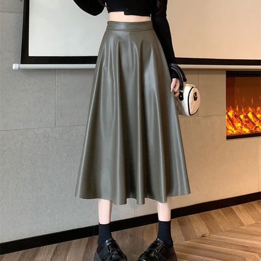 Frau in schwarzem Oberteil und langem, olivgrünem Lederrock, steht vor einem Kamin. Modetrend, eleganter Look, Herbstmode.