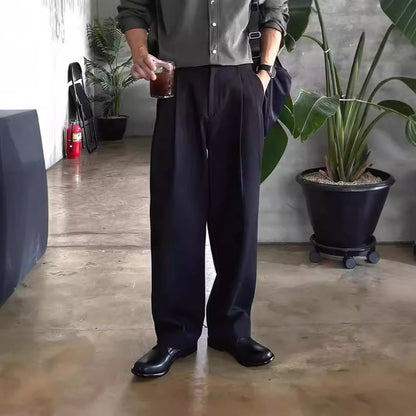 Mann in schwarzer Hose und Hemd hält Getränk, steht neben Pflanze in modernem Raum. Elegante Herrenmode, lässiger Stil, Indoor-Pflanzen.