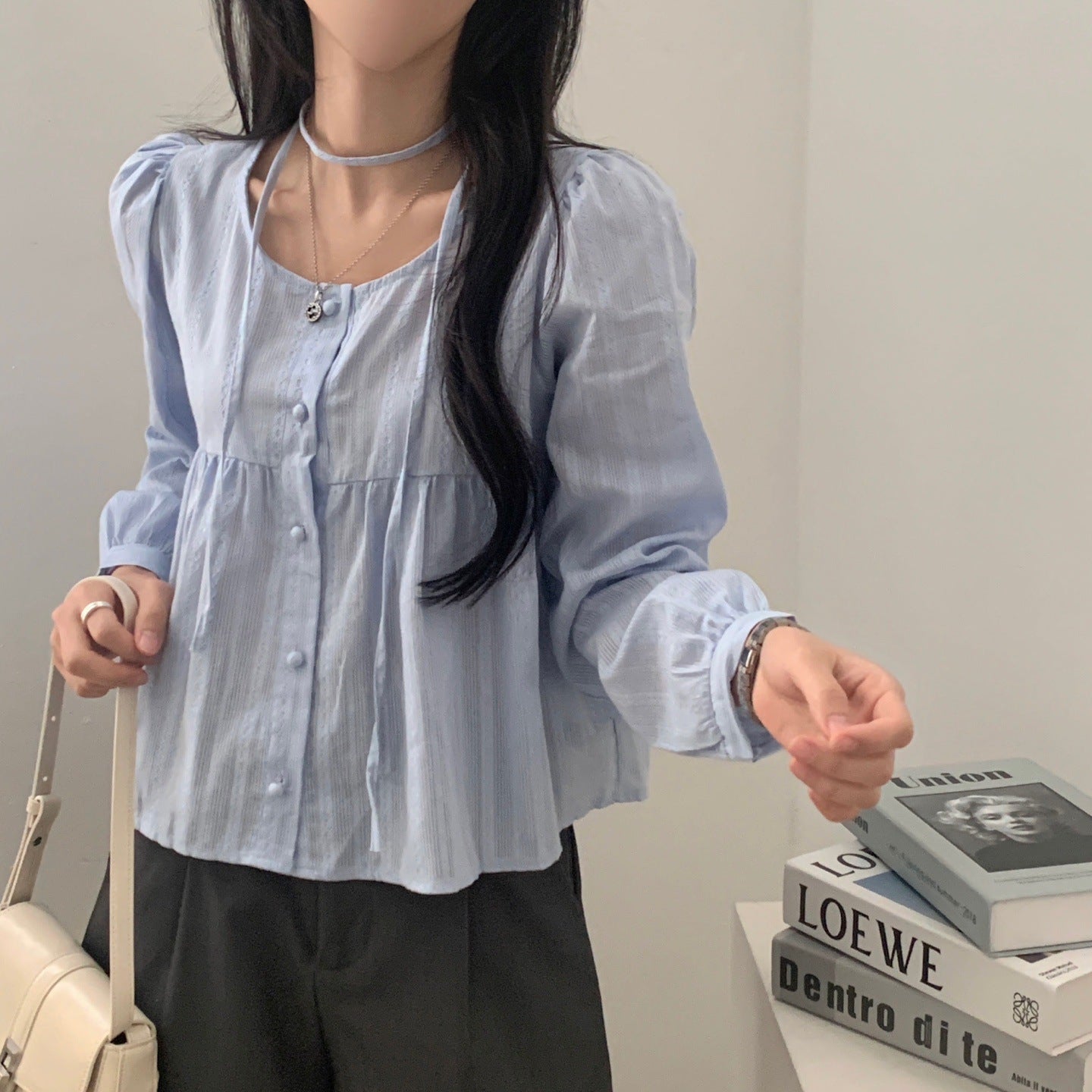 Frau in hellblauer Bluse mit langen Ärmeln, hält weiße Handtasche. Modetrend, elegante Damenbekleidung, stilvolles Outfit, Freizeitlook.