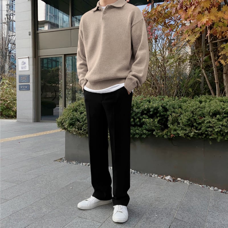 Ein Mann in beiger Strickjacke und schwarzer Hose steht vor einem modernen Gebäude. Herbstliche Pflanzen im Hintergrund, weiße Sneakers.