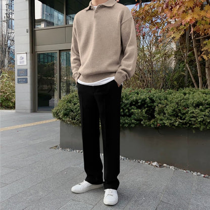 Ein Mann in beiger Strickjacke und schwarzer Hose steht vor einem modernen Gebäude. Herbstliche Pflanzen im Hintergrund, weiße Sneakers.