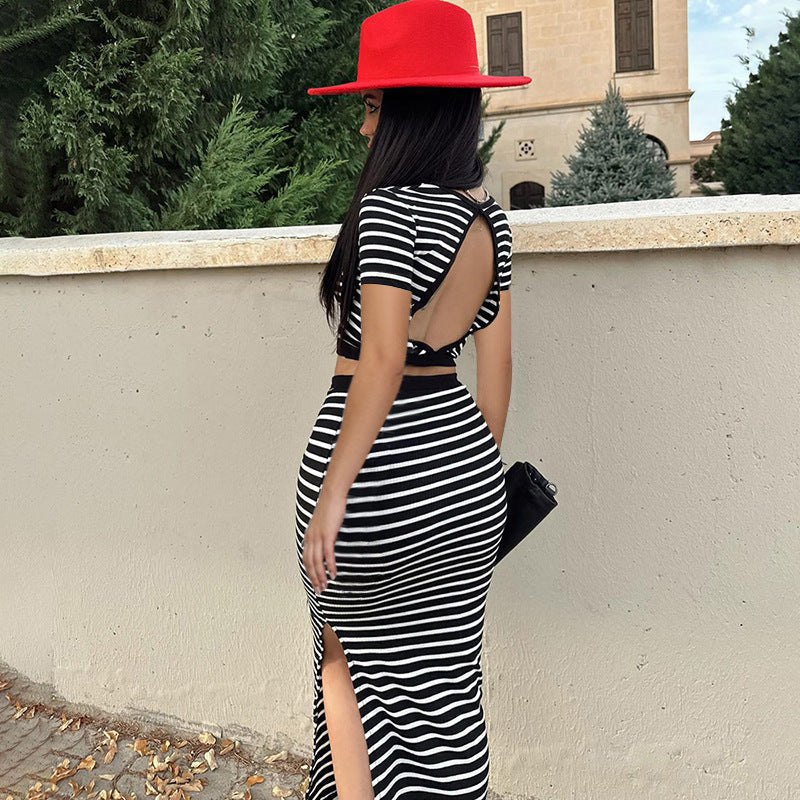 Frau in gestreiftem Kleid mit Rückenausschnitt und rotem Hut steht vor einer Mauer, umgeben von Bäumen. Elegante Mode, Sommeroutfit, Streetstyle.