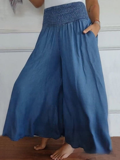 Frau trägt weite, blaue Palazzo-Hose mit elastischem Bund, barfuß auf Holzboden. Modetrend, bequeme Damenbekleidung, lässiger Sommerstil.