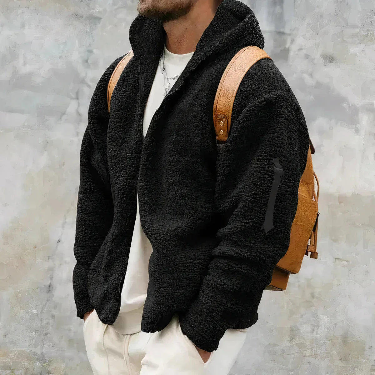 Mann in schwarzer Fleecejacke mit Kapuze und braunem Rucksack vor grauem Hintergrund. Modetrend, Herrenbekleidung, Wintermode.