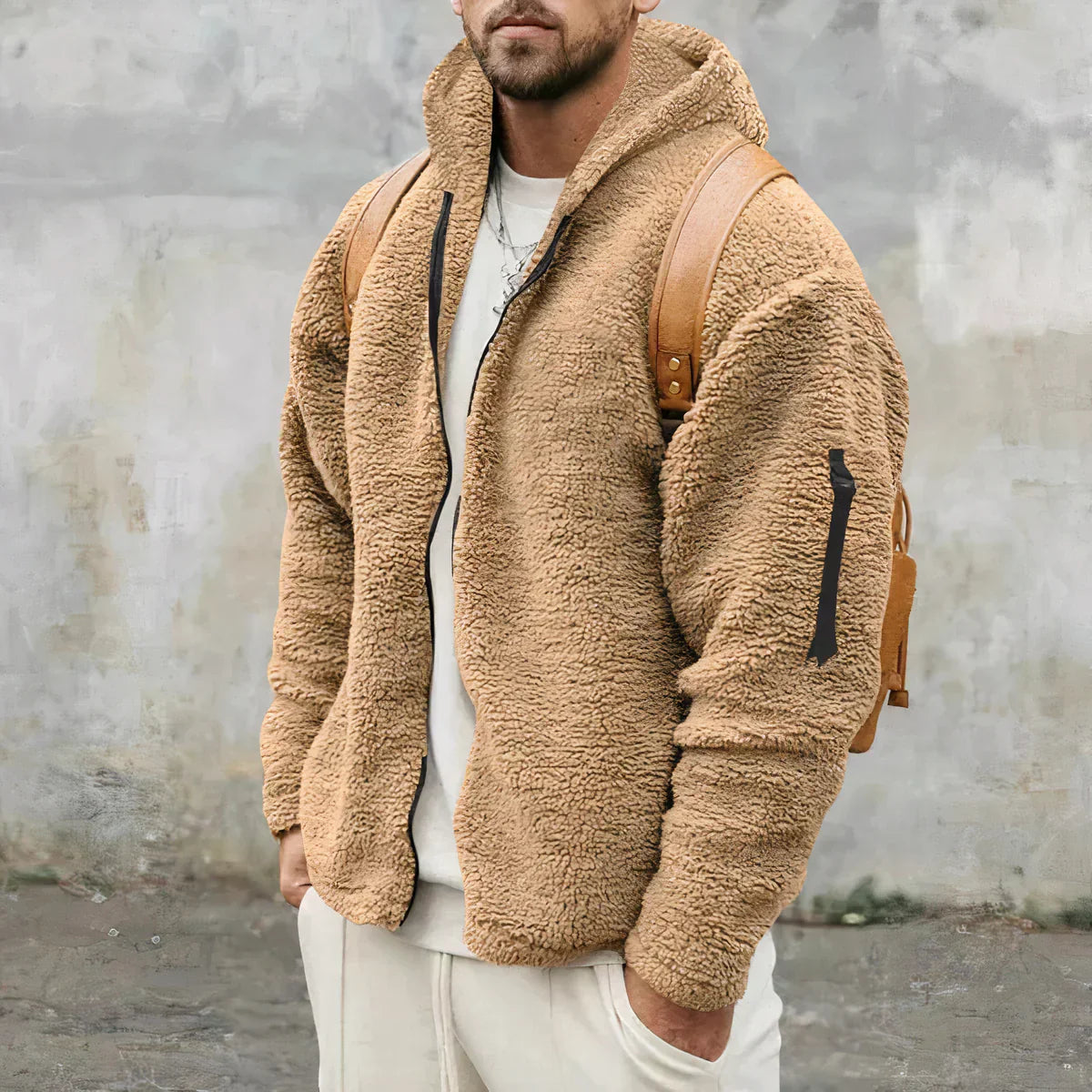 Mann in beiger Teddyfelljacke mit Kapuze und braunem Rucksack vor grauer Wand. Modetrend, Winterjacke, Herrenmode, Outdoor-Bekleidung.