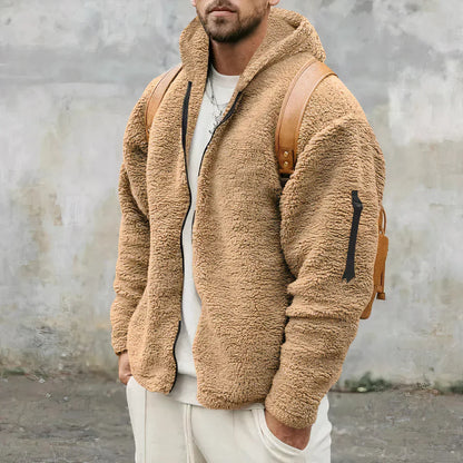 Mann in beiger Teddyfelljacke mit Kapuze und braunem Rucksack vor grauer Wand. Modetrend, Winterjacke, Herrenmode, Outdoor-Bekleidung.