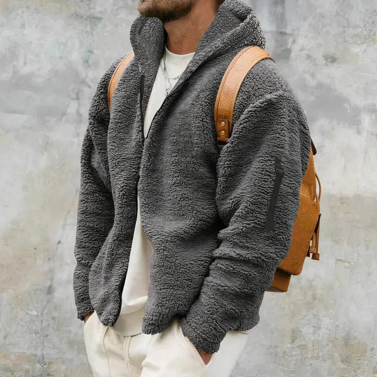 Mann in grauer Fleecejacke mit braunem Rucksack vor grauem Hintergrund. Modetrend, Herrenbekleidung, Winterjacke, lässiger Stil.