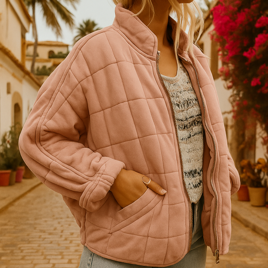 Frau in rosa Steppjacke auf gepflasterter Straße mit Palmen und blühenden Bougainvillea im Hintergrund. Modetrend, Herbstmode, Outdoor-Kleidung.