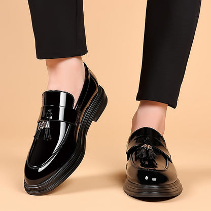 Schwarze Lackleder-Loafer mit Quasten, getragen von einer Person in schwarzer Hose, auf beigem Hintergrund. Elegante Herrenschuhe, modisch und glänzend.