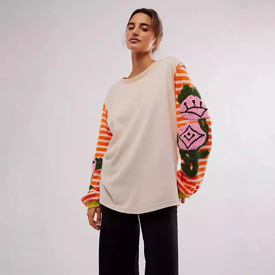 Frau trägt beige Pullover mit bunten, gestreiften Ärmeln und Blumenmuster, kombiniert mit schwarzen Hosen. Modetrend, Damenmode, Herbstoutfit.
