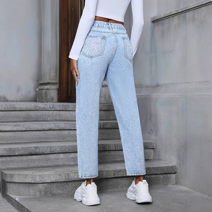 Frau in hellblauer Mom-Jeans mit Stickerei, weißem Langarmshirt und weißen Sneakers auf Treppe. Modetrend, lässiger Streetstyle.