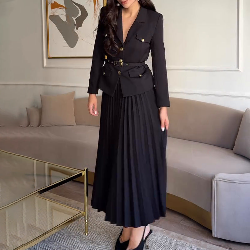 Elegante Frau in schwarzem Blazer und plissiertem Rock, steht in modernem Wohnzimmer mit beigem Sofa und abstraktem Kunstwerk an der Wand.