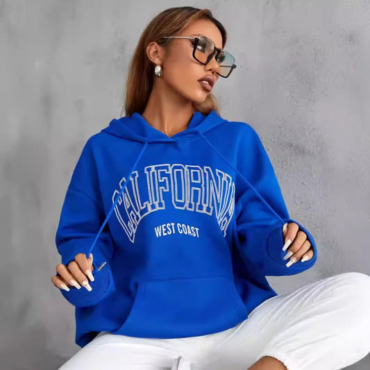 Frau in blauem "California West Coast" Hoodie, trägt Sonnenbrille, sitzt lässig vor grauer Wand. Modetrend, Freizeitkleidung, Streetwear.