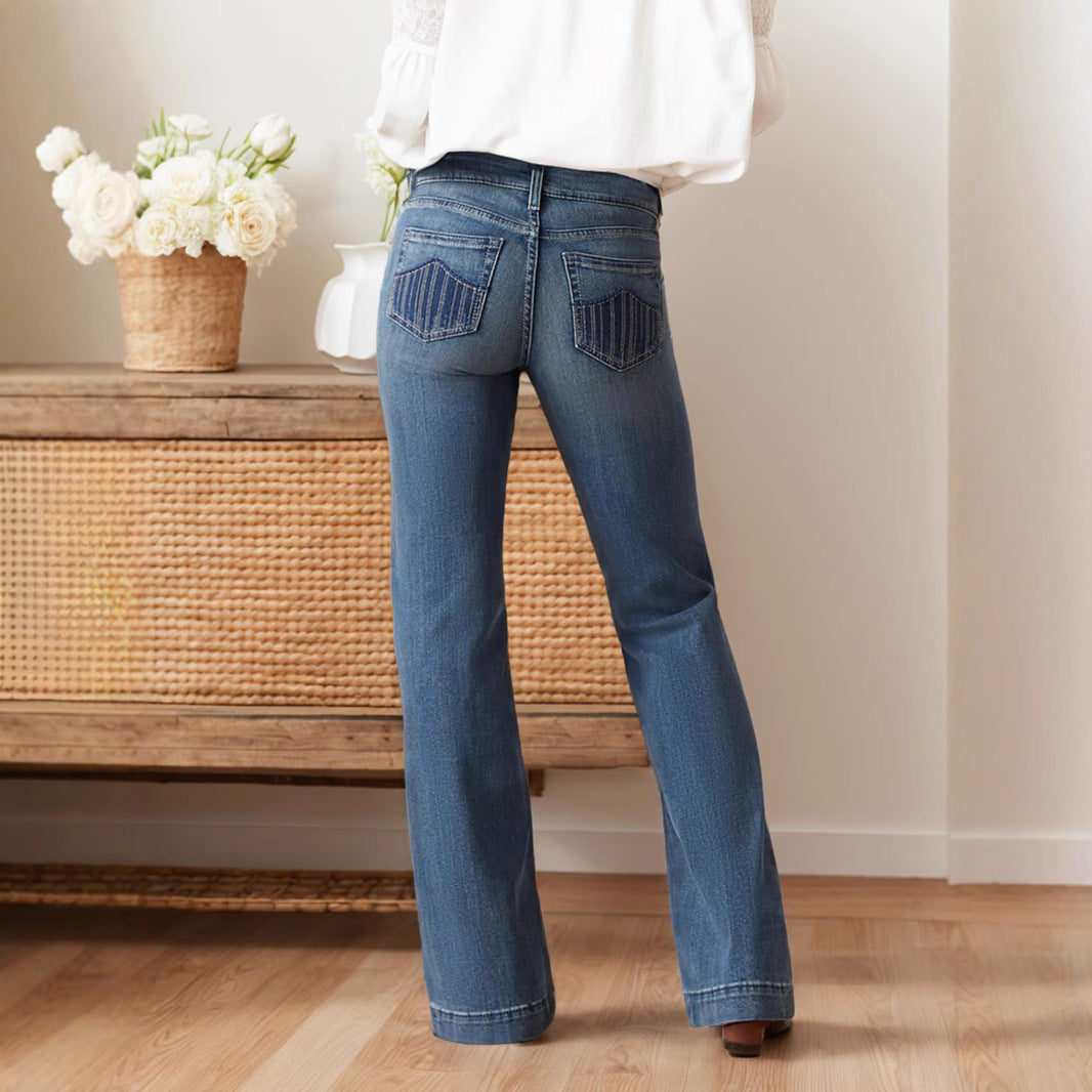 Frau in blauer Schlagjeans steht vor einem Holztisch mit Blumenkorb, helle Wand im Hintergrund. Modetrend, Damenjeans, Freizeitkleidung.
