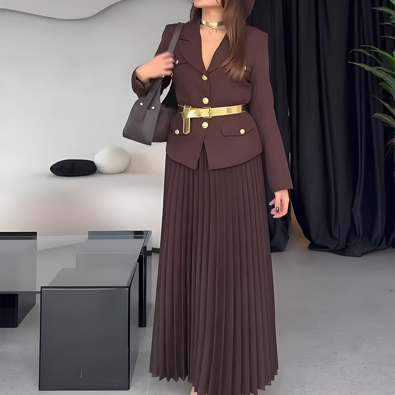 Elegante Frau in braunem Anzug mit goldener Gürtelschnalle, langer Plissee-Rock, schwarze Handtasche, modernes Interieur, stilvolle Mode.