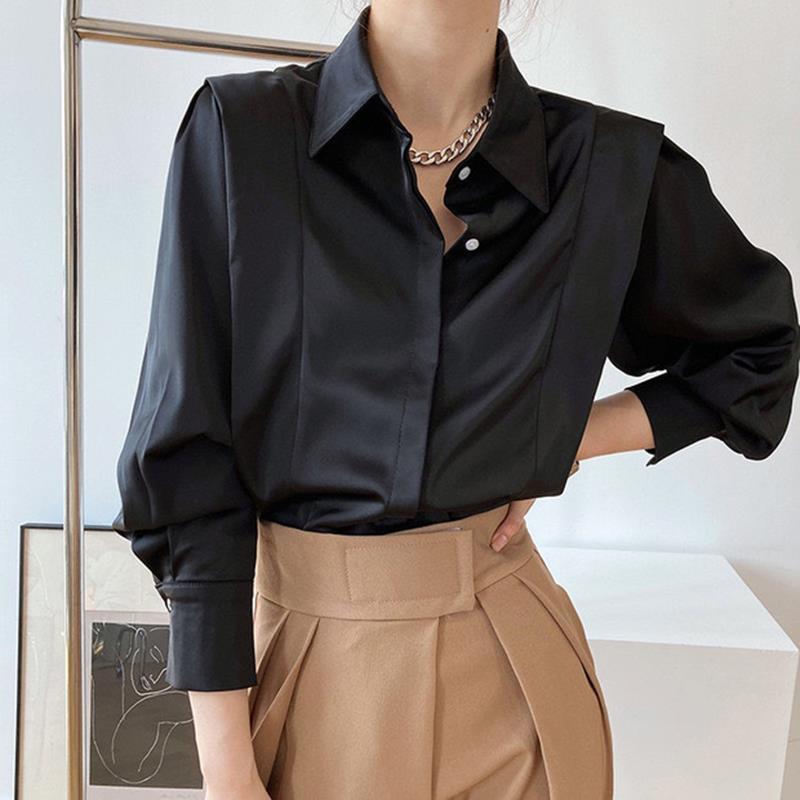 Frau in schwarzer Satinbluse und beigen High-Waist-Hosen, modisch elegant, mit Kette, vor minimalistischem Hintergrund.