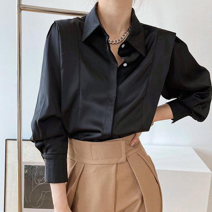 Frau in schwarzer Satinbluse und beigen High-Waist-Hosen, modisch elegant, mit Kette, vor minimalistischem Hintergrund.