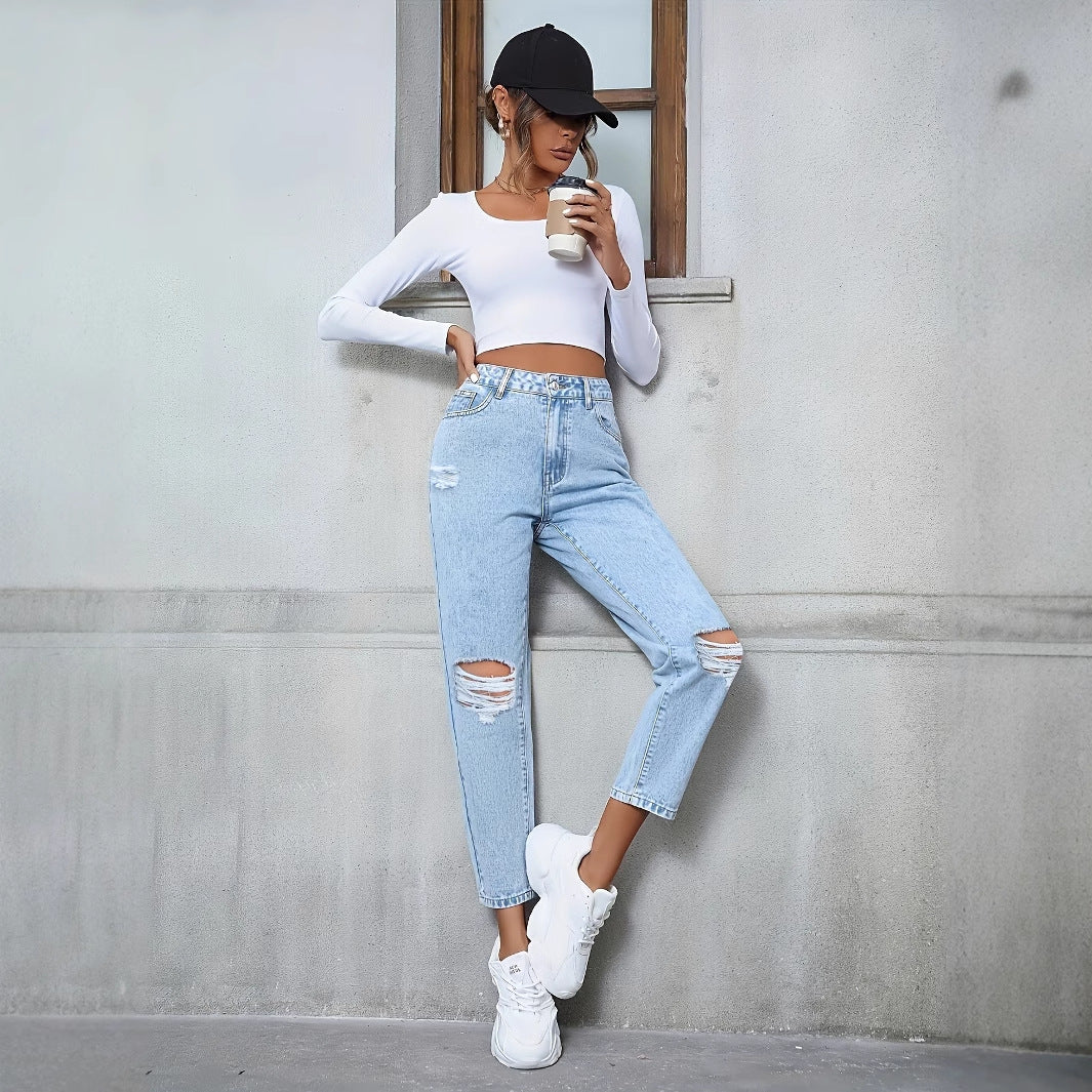 Frau in zerrissener Jeans und weißem Crop-Top lehnt an Wand, hält Kaffeebecher, trägt schwarze Kappe und weiße Sneaker. Urbaner Streetstyle-Look.