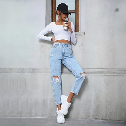 Frau in zerrissener Jeans und weißem Crop-Top lehnt an Wand, hält Kaffeebecher, trägt schwarze Kappe und weiße Sneaker. Urbaner Streetstyle-Look.