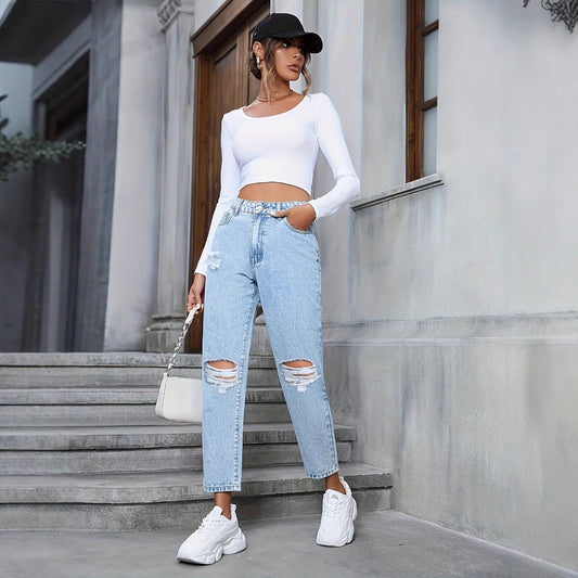 Frau in weißem Crop-Top und zerrissenen Jeans, trägt weiße Sneaker und schwarze Kappe, steht vor grauem Gebäude. Modetrend, Streetstyle, lässig.